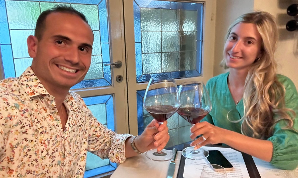 A couple toasting with glasses of red wine during a cozy dinner by a stained glass window.