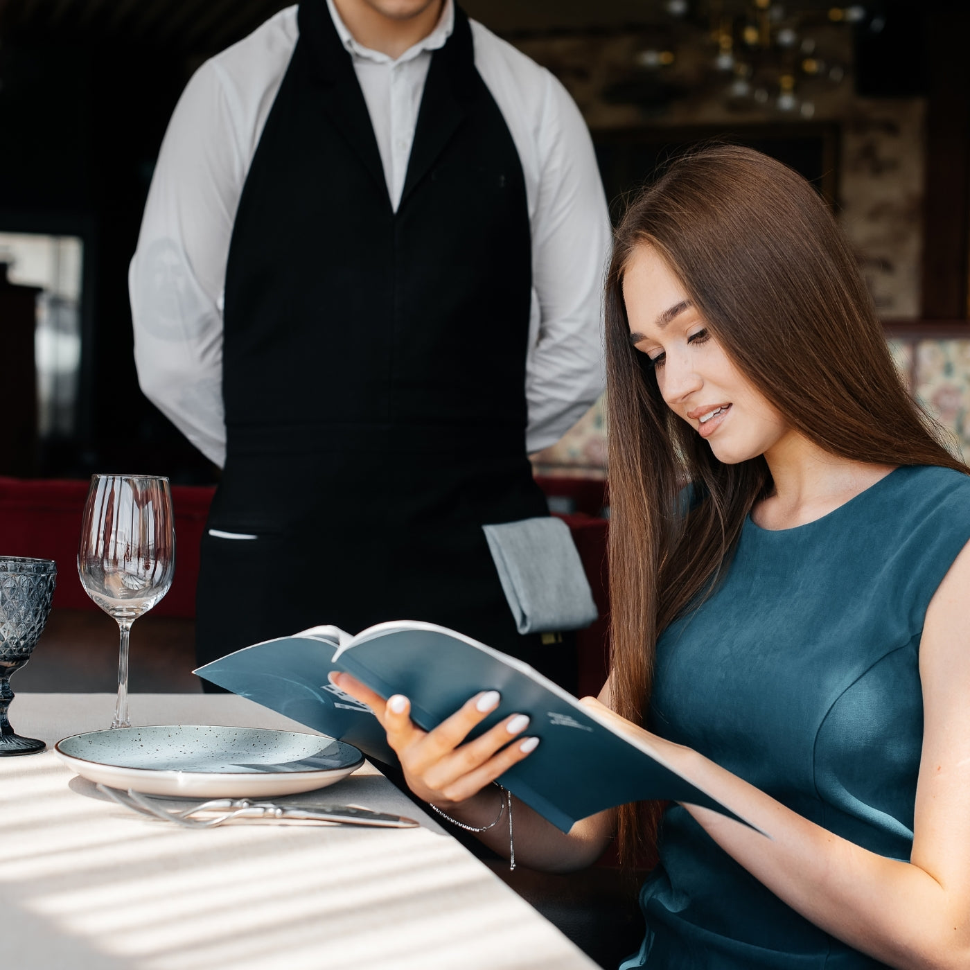 A young woman in an elegant restaurant browsing the menu and placing an order with a waiter in a stylish apron. Fine dining and excellent customer service.