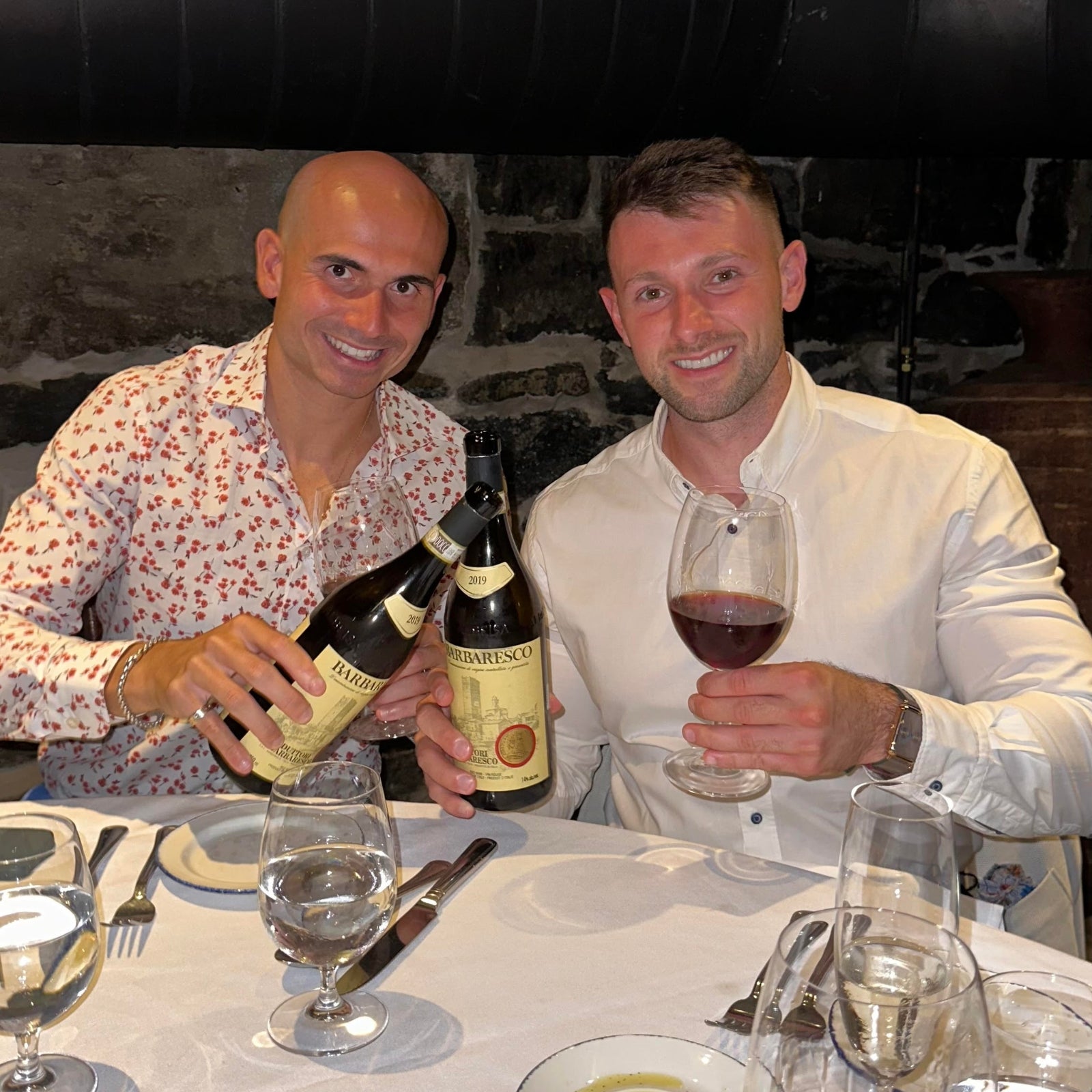 Two friends holding bottles of Barbaresco wine and enjoying a dinner with red wine at a restaurant.
