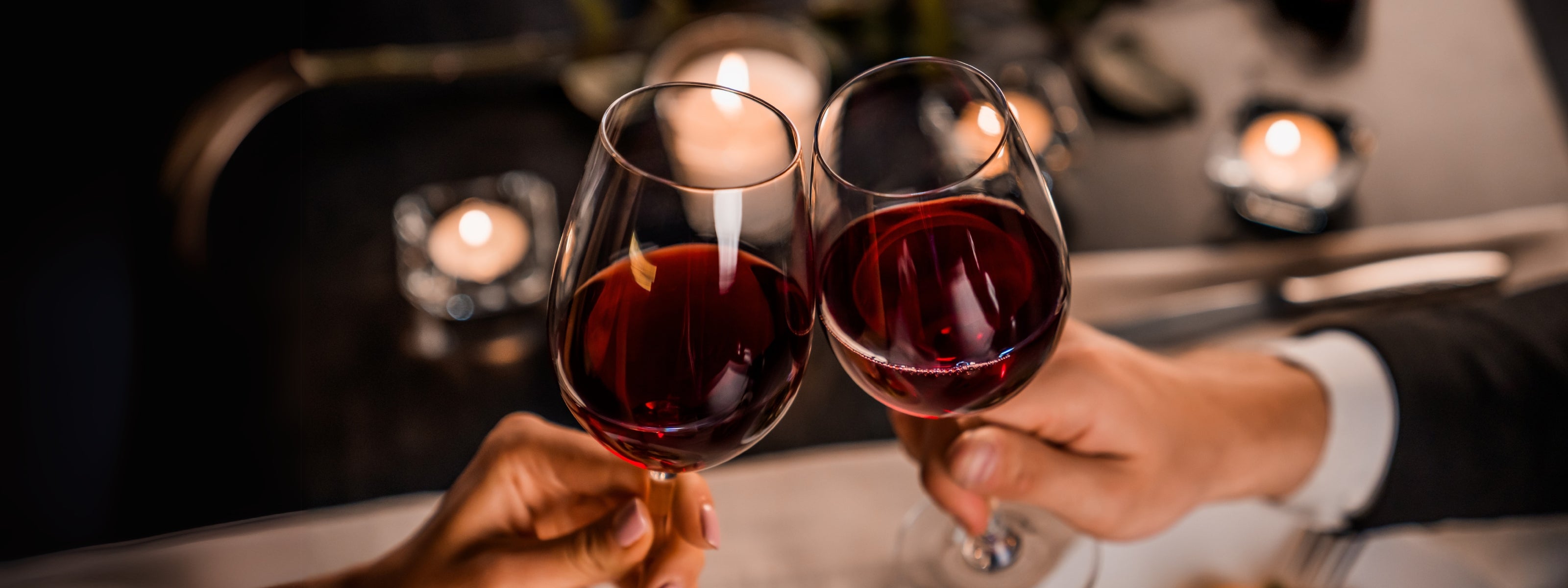 A close-up of two hands toasting glasses of red wine in a romantic candlelit setting, creating a warm and intimate atmosphere.