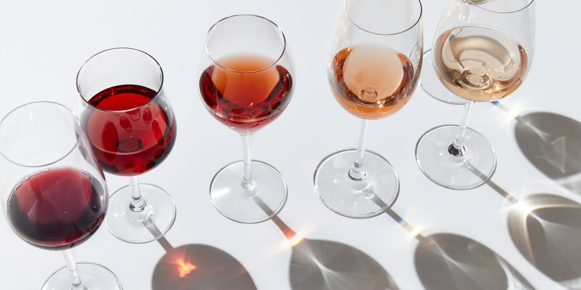 A line of wine glasses filled with red, white, and rosé wine on a white background, with reflections and shadows.