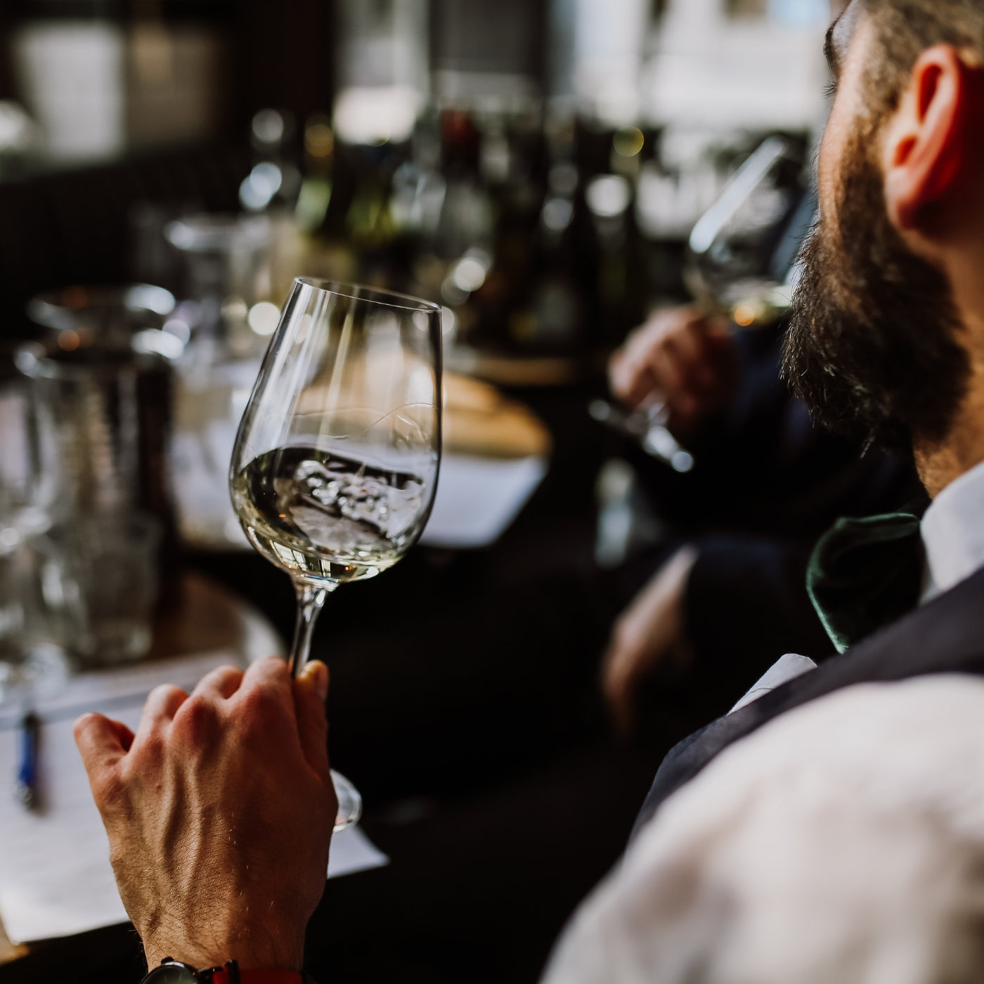 A young European man holding a glass of white wine during a wine tasting session. The focus is on the wine glass and the man, with bottles and glasses blurred in the background.